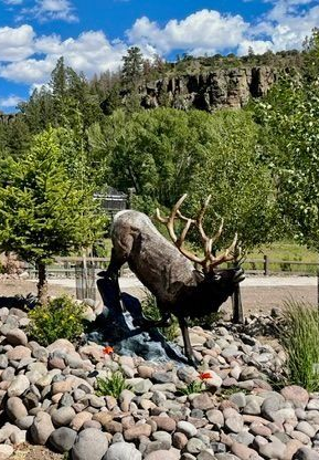 Bronze elk statue in a rock garden with trees and a rocky cliff under a blue sky
