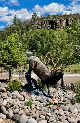 Bronze elk statue in a rock garden with trees and a rocky cliff under a blue sky