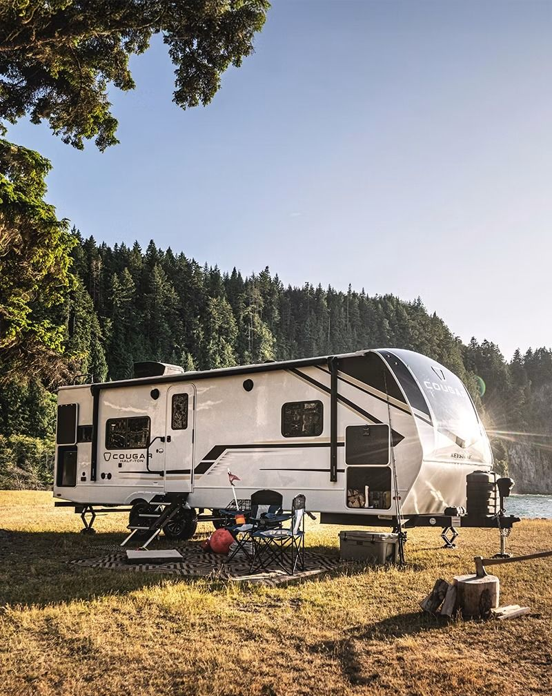 White travel trailer parked by a lakeshore with camping chairs and a fire pit in a wooded area