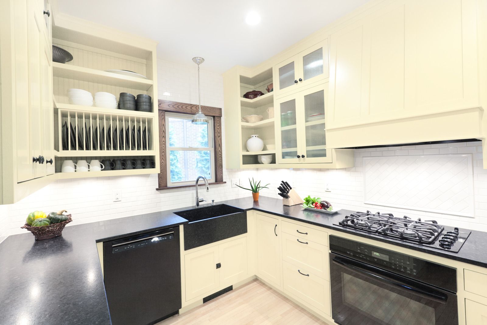 Cream-colored kitchen with black countertops, appliances. Plate rack, glass-front cabinets, farmhouse sink, and gas cooktop.