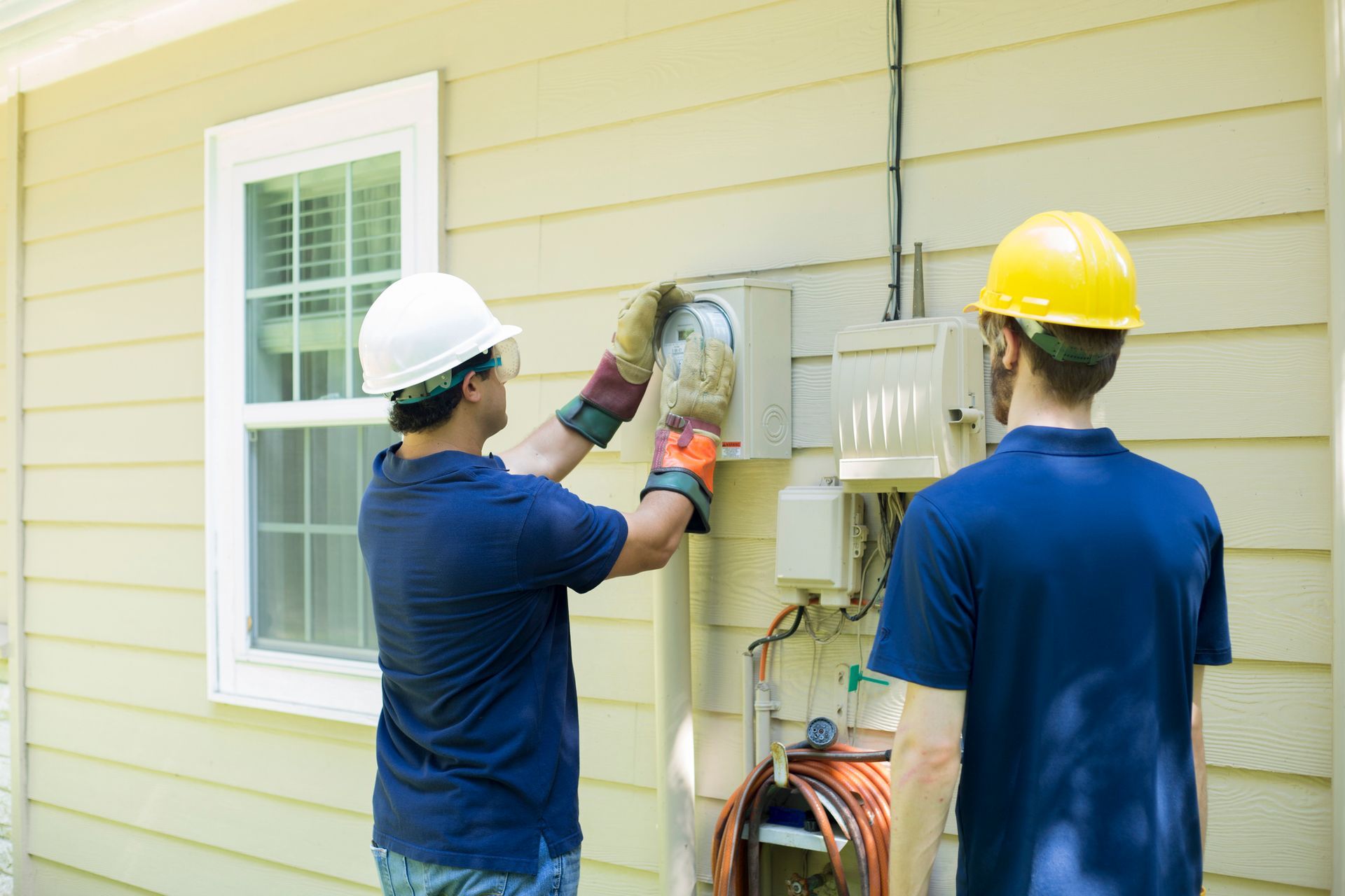 Two men are working on a meter on the side of a house. Two men are working on a meter on the side of a house.