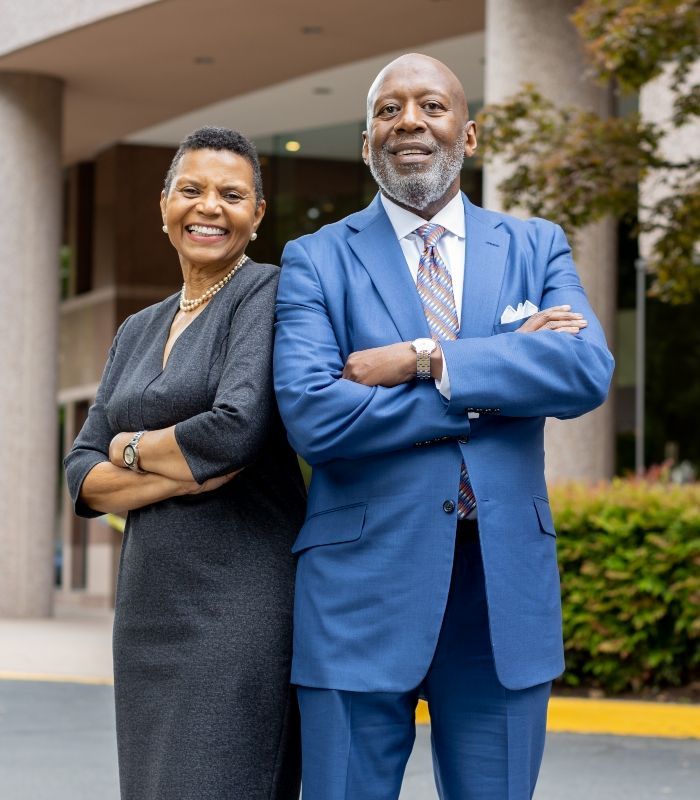 Man and woman standing with arms crossed; building exterior. Man in blue suit, woman in gray dress, both smiling.