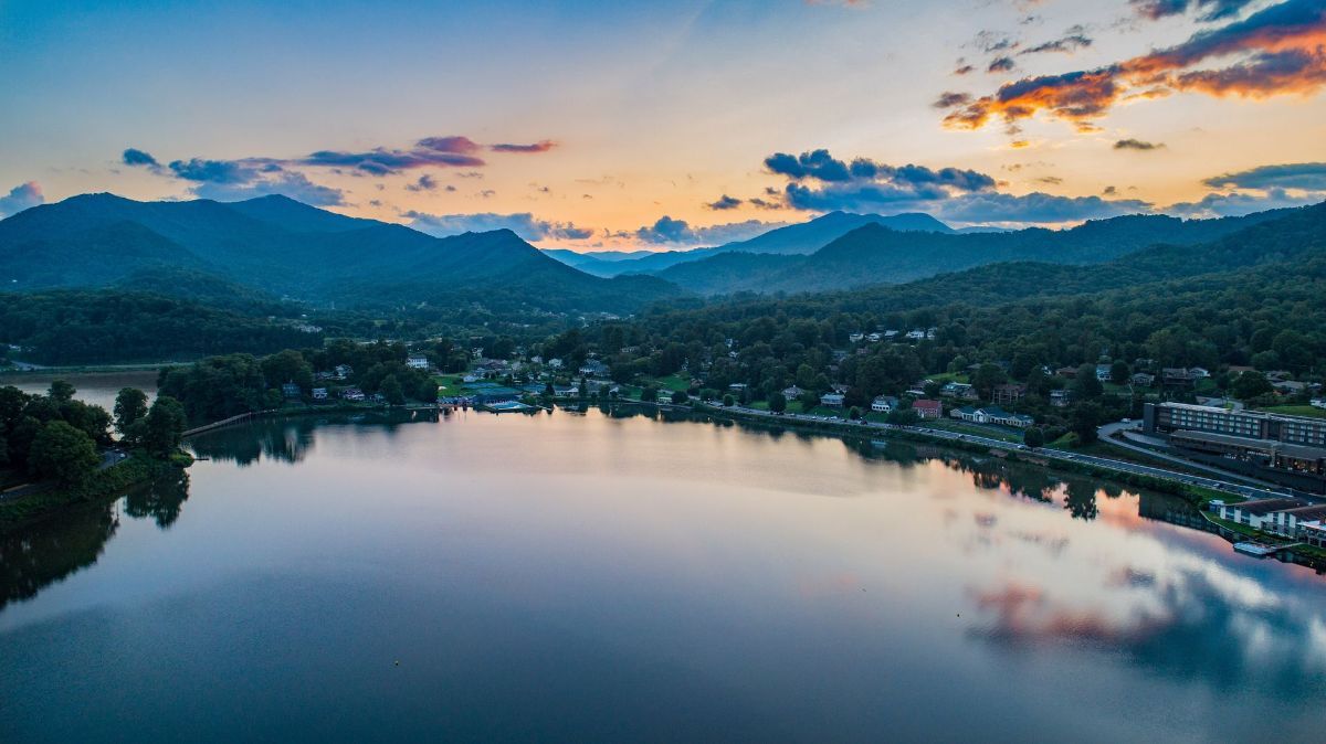 A wide, calm lake reflecting a vibrant orange and purple sunset beneath a mountain range and a small town.