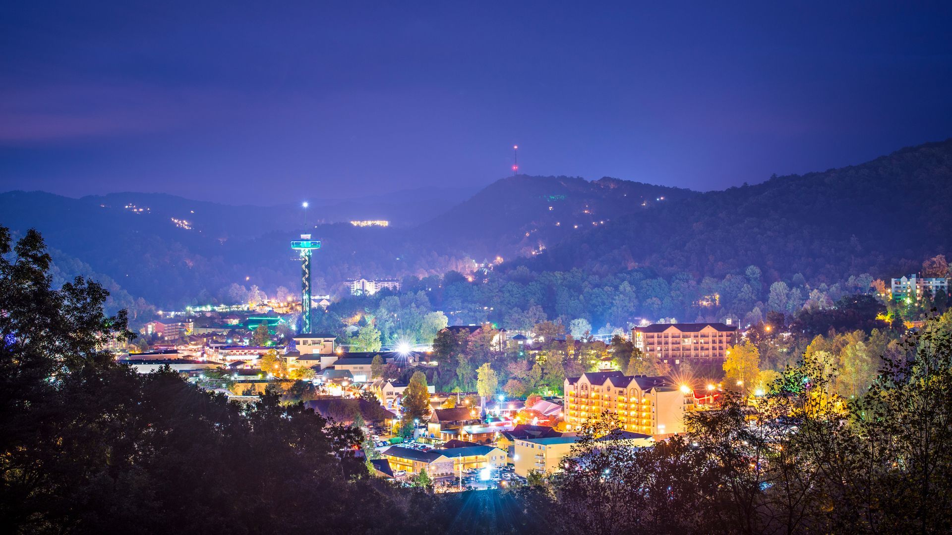 Nighttime cityscape with colorful lights nestled among hills under a purple-blue sky