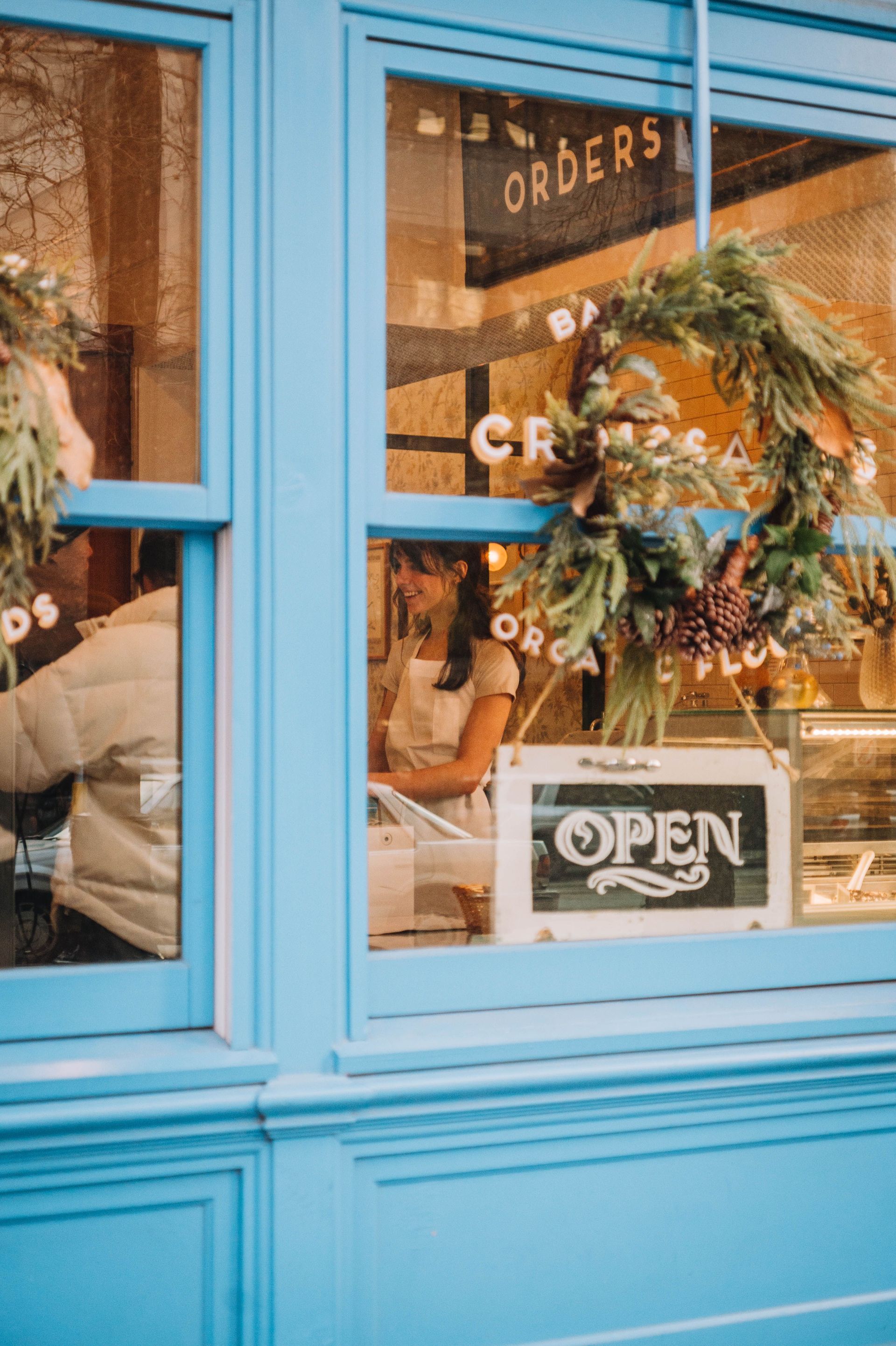 Blue storefront window with an “OPEN” sign, wreath, and flower arrangements