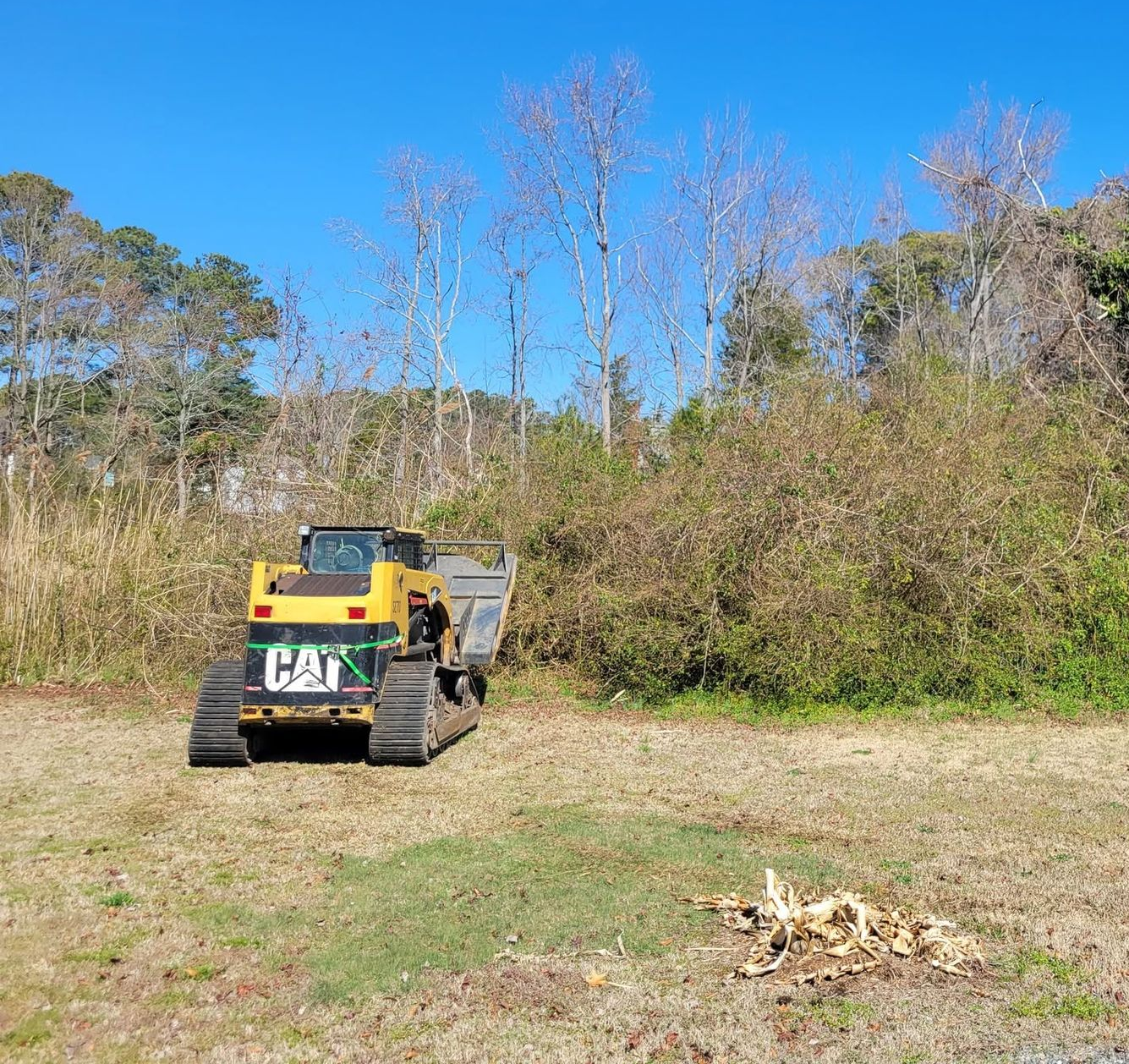 Yellow tracked utility vehicle on a grassy field beside trees under a clear blue sky