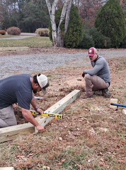 Two workers measuring a wooden beam outdoors with a tape measure and level in a leaf-covered yard