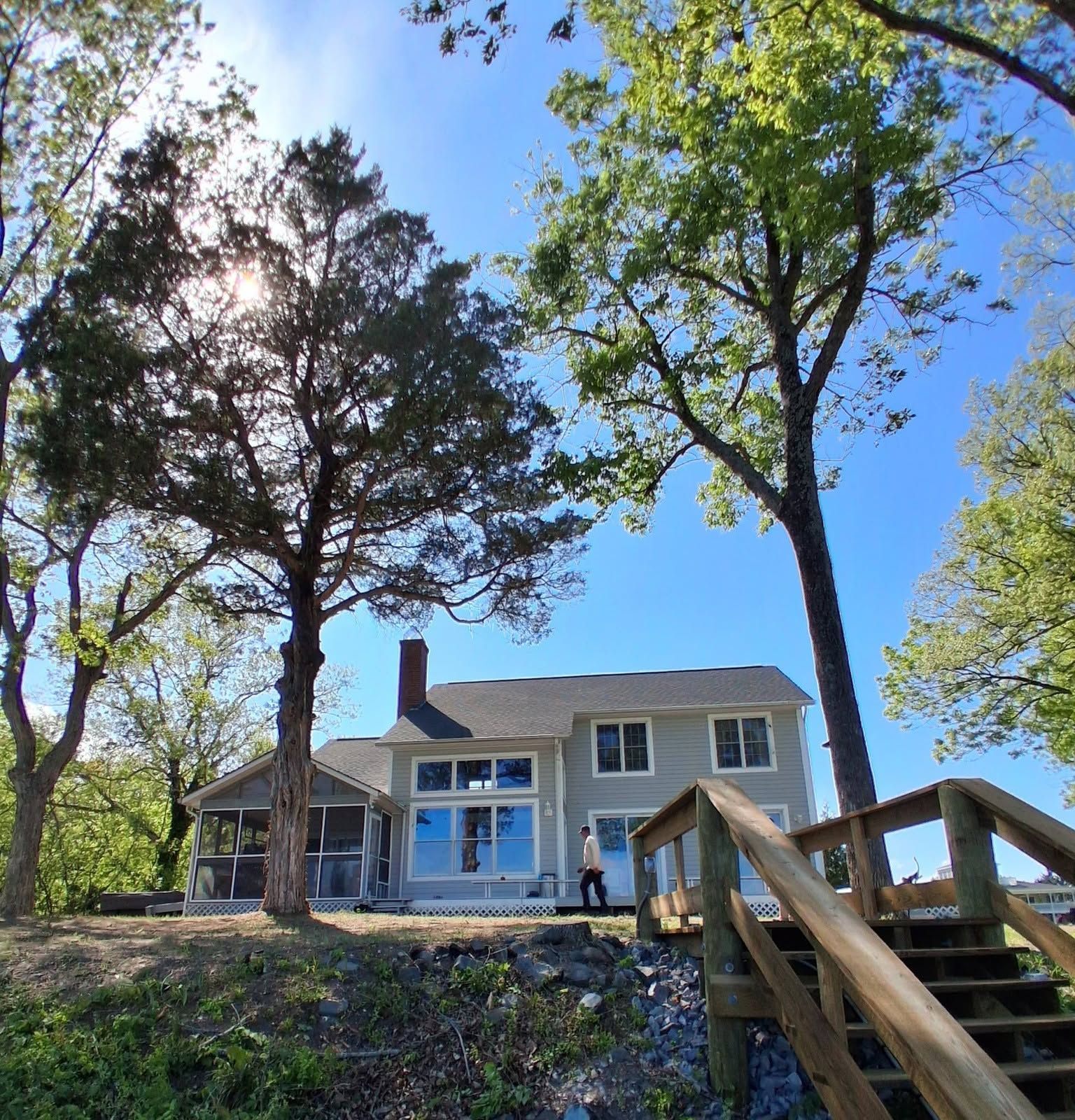 Two-story house on a wooded hillside with stairs and trees under a bright blue sky