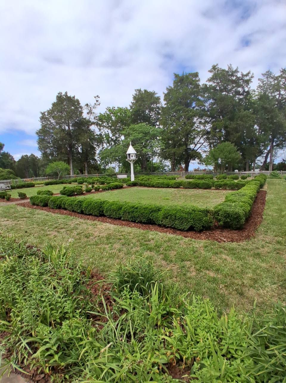 Formal garden with trimmed hedges, grassy paths, and a white lamp post under a cloudy sky