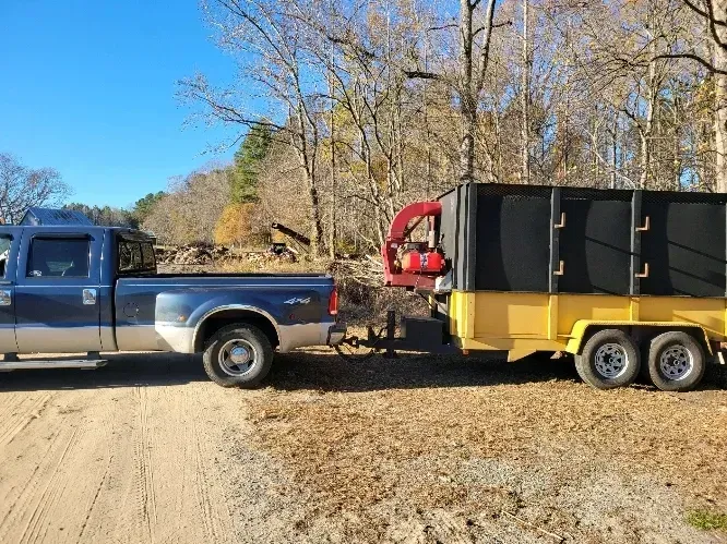 Blue pickup truck towing a yellow dump trailer with a red machine on a dirt road in a wooded area