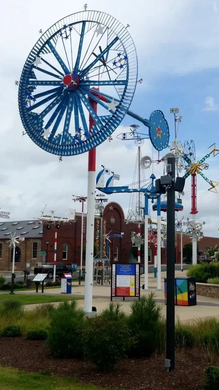 Large blue decorative bicycle sculpture in a public park with flowers, trees, and brick buildings behind it