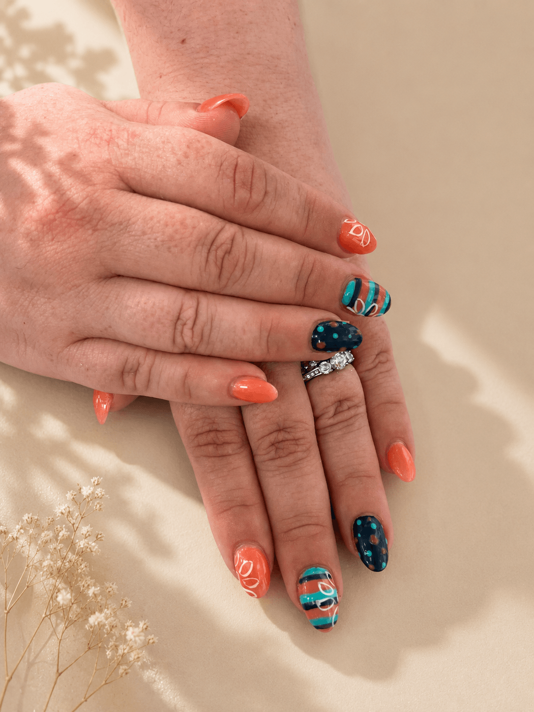 Hands with coral and turquoise nail art resting on a beige surface.