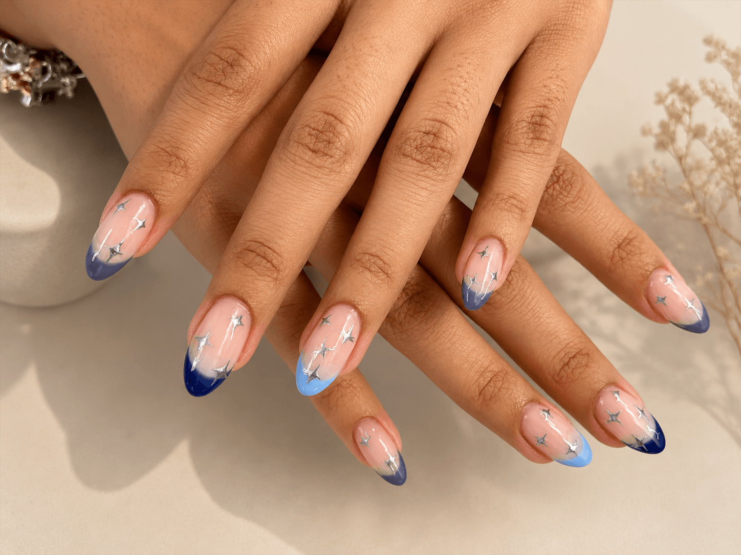 Hands with long nude nails tipped in blue, resting on a beige surface.