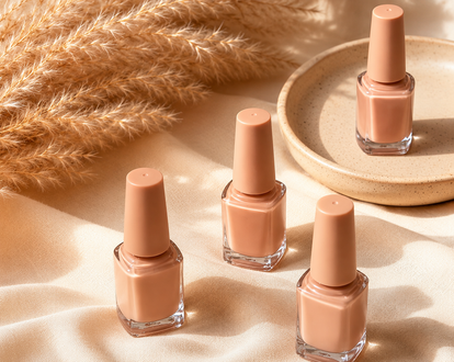 Four nude nail polish bottles on a soft beige fabric backdrop with dried pampas grass and a tray.