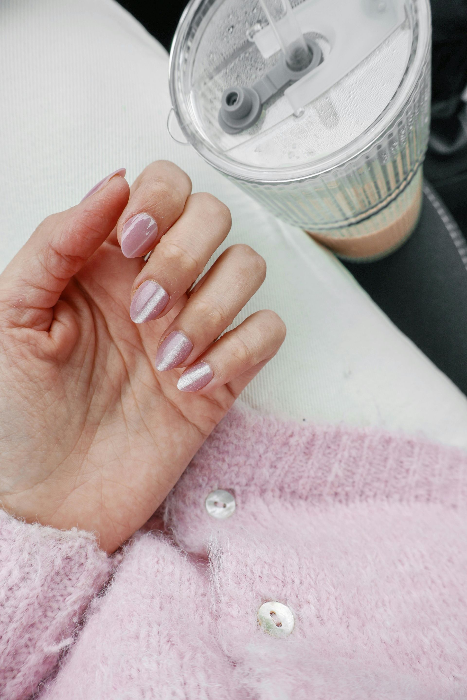 Hand holding an iced drink beside a pink textured sweater with pearl buttons.