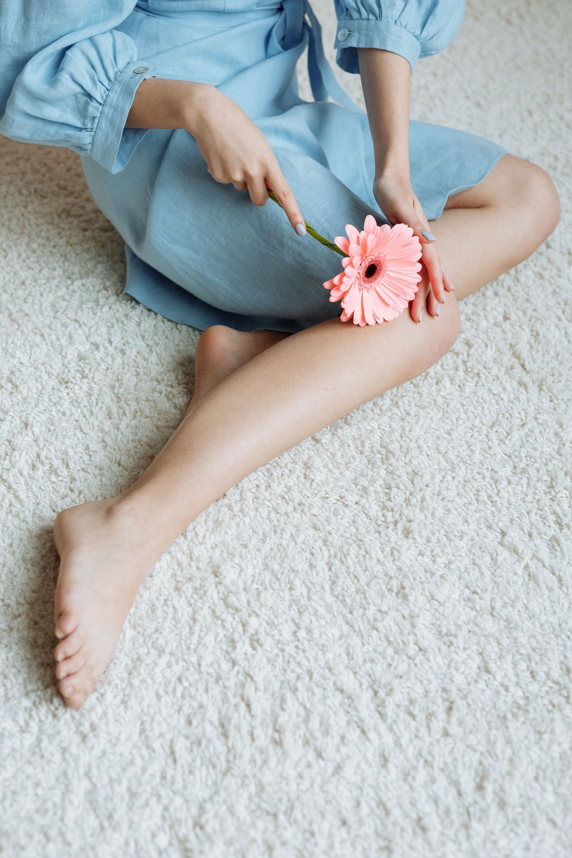 Person in a blue dress sitting on a white rug, holding a pink flower against a bent knee.