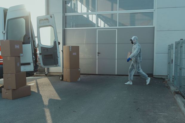 Person in white protective suit walking near stacked cardboard boxes outside a warehouse.