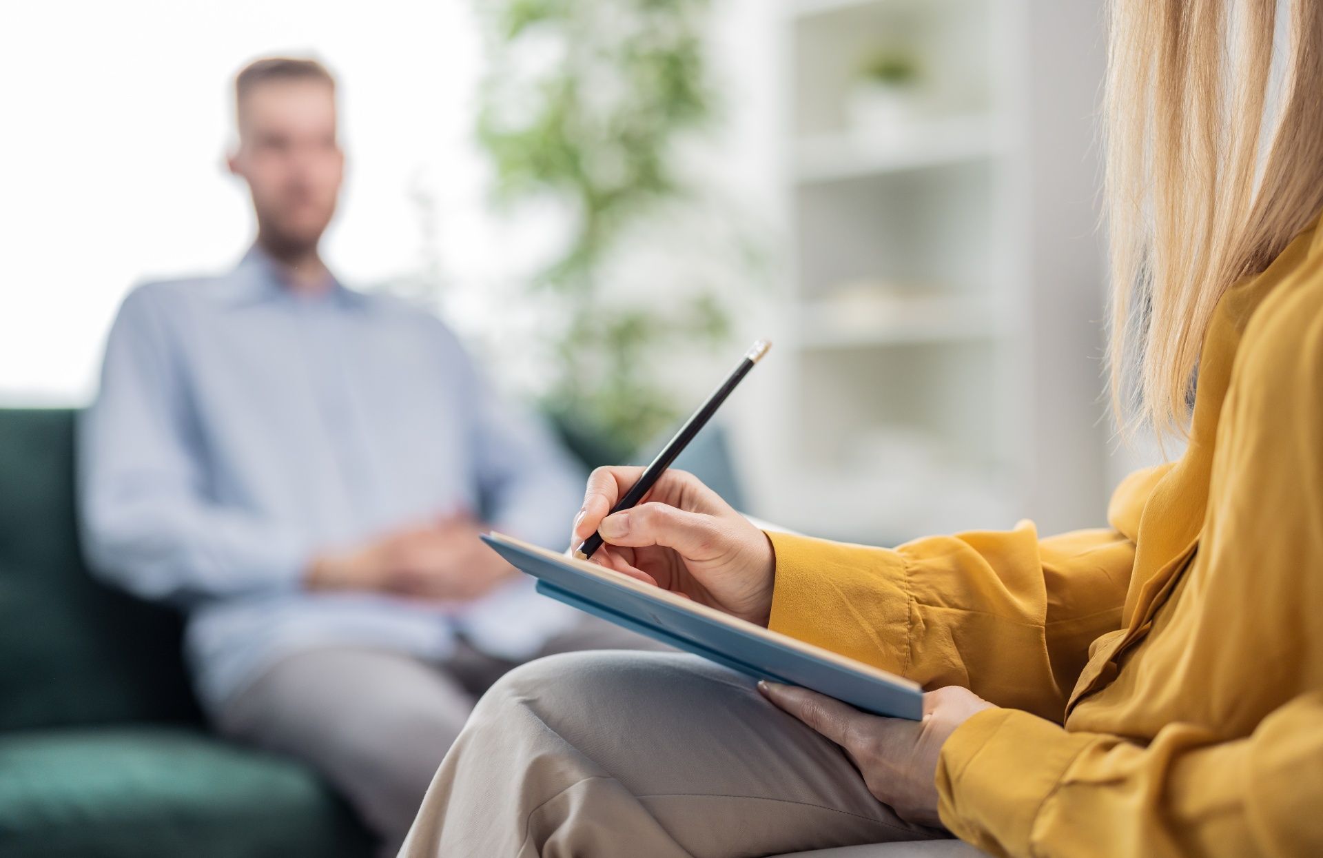A woman is sitting on a couch writing in a notebook while a man sits in the background.