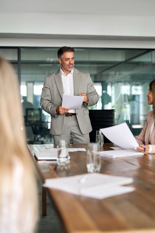Business meeting in a glass-walled conference room, with a man presenting papers to seated colleagues.