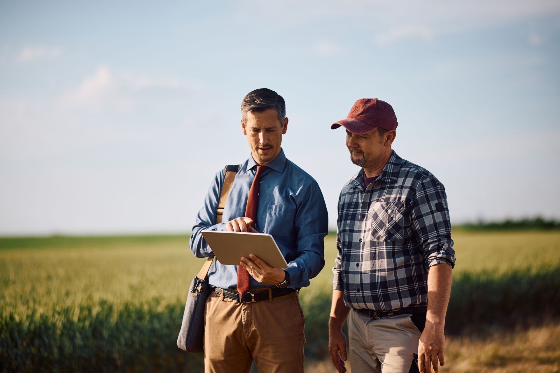 Two farmers in a field reviewing a tablet, one pointing at the screen under a bright sky