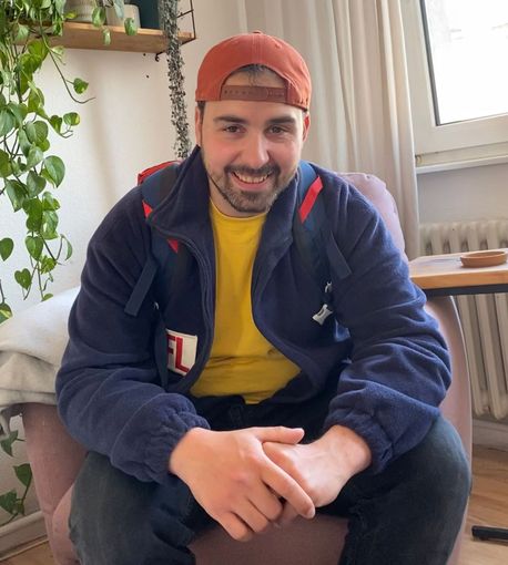 Elektroniker Torsten Wiland mit orangefarbener Mütze und blauer Jacke sitzt auf einem Stuhl und lächelt freundlich und professionell in die Kamera.