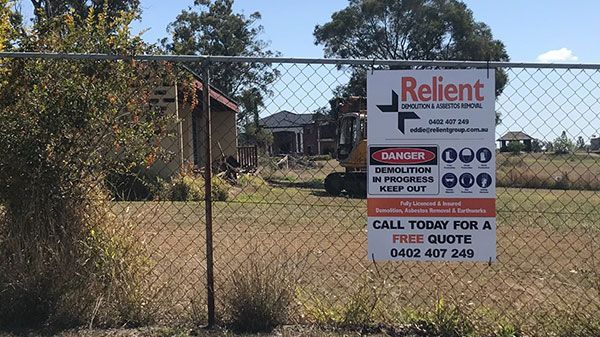 A yellow excavator is demolishing a house behind a fence.