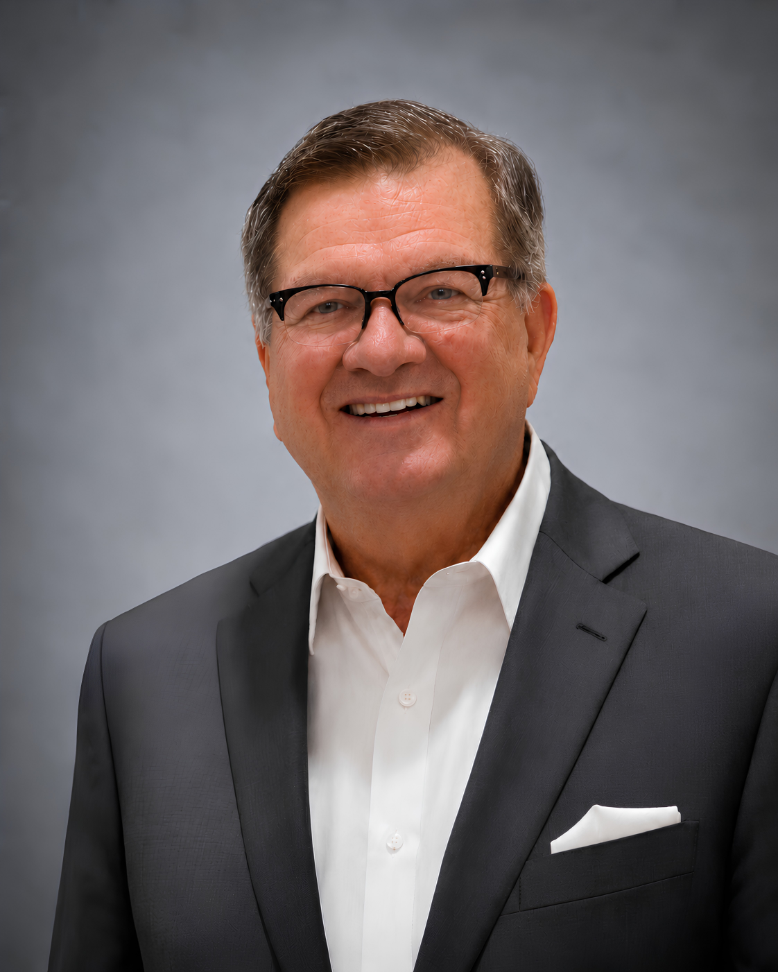 Studio headshot of a smiling man in a dark suit, white shirt, and glasses against a gray background