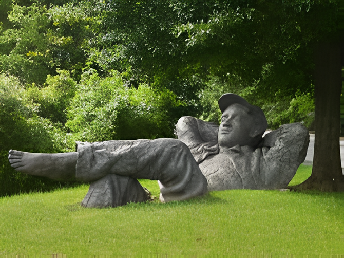 Large reclining stone statue in a grassy park under trees, with one arm behind its head.