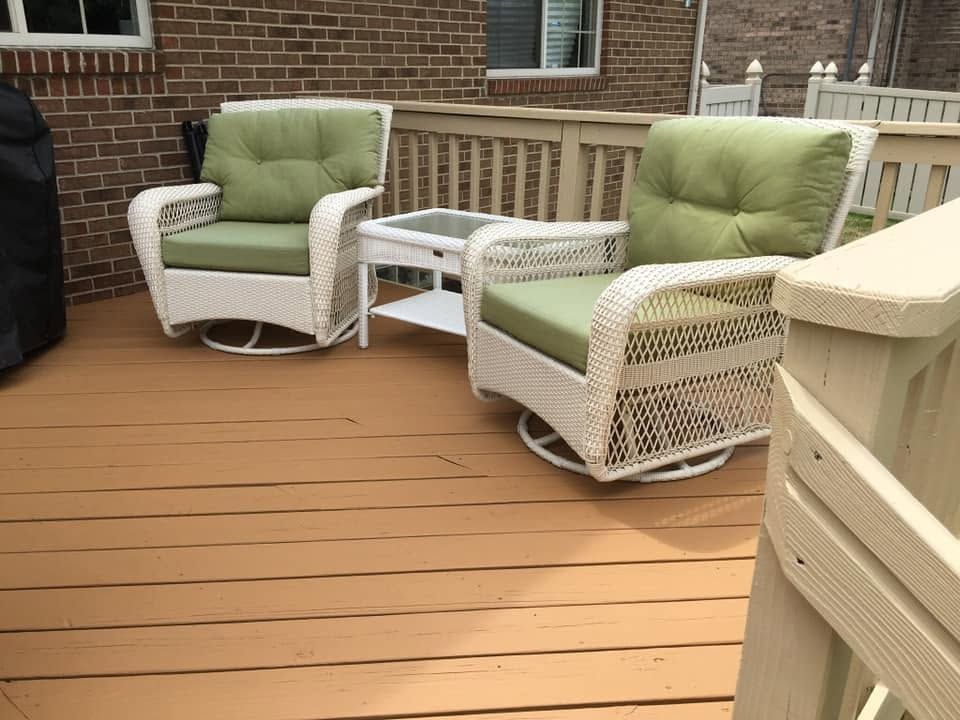 Two green cushioned wicker chairs on a wooden deck with a small table against a brick house wall