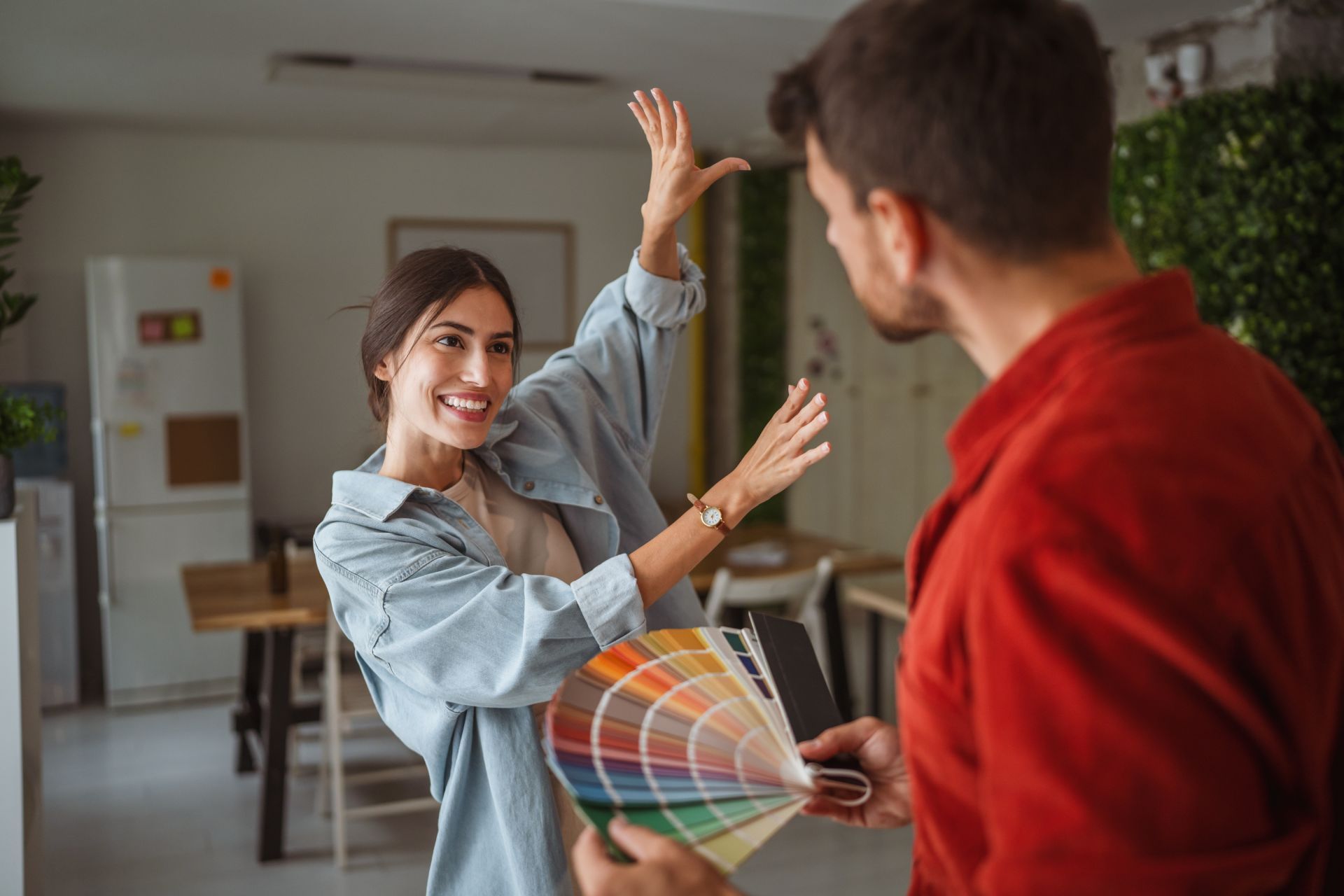 Two coworkers in a bright office, smiling and holding color swatches while one raises a hand.