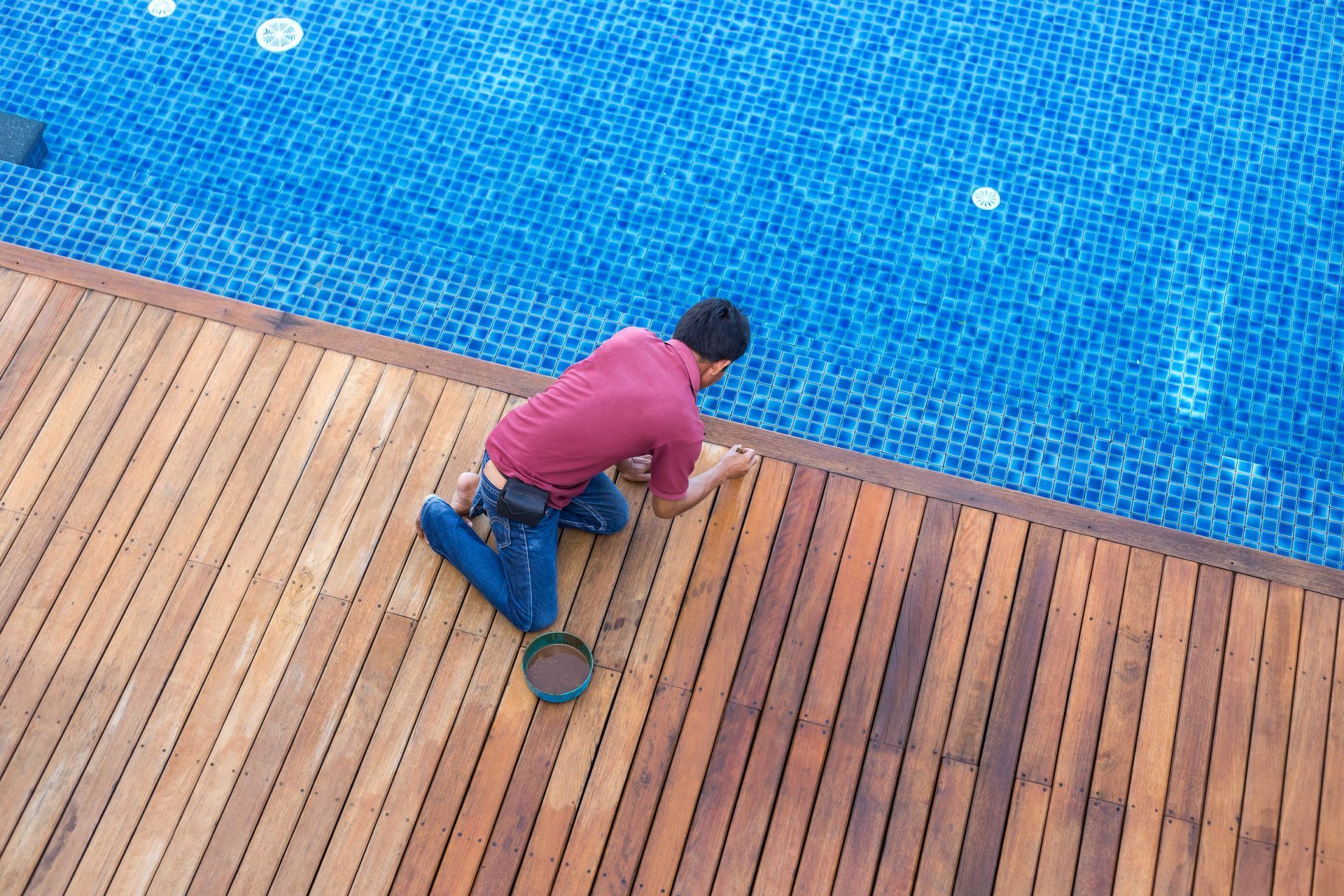 Person kneels on a wooden deck beside a blue swimming pool, reaching toward a tennis racket.