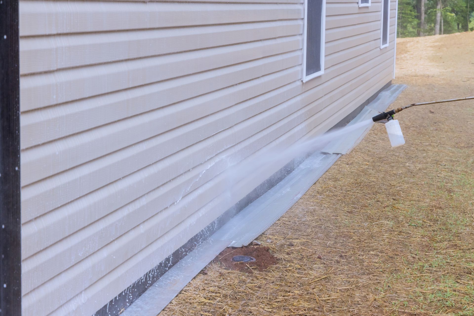 House siding with plastic moisture barrier exposed along the foundation beside a gravel driveway
