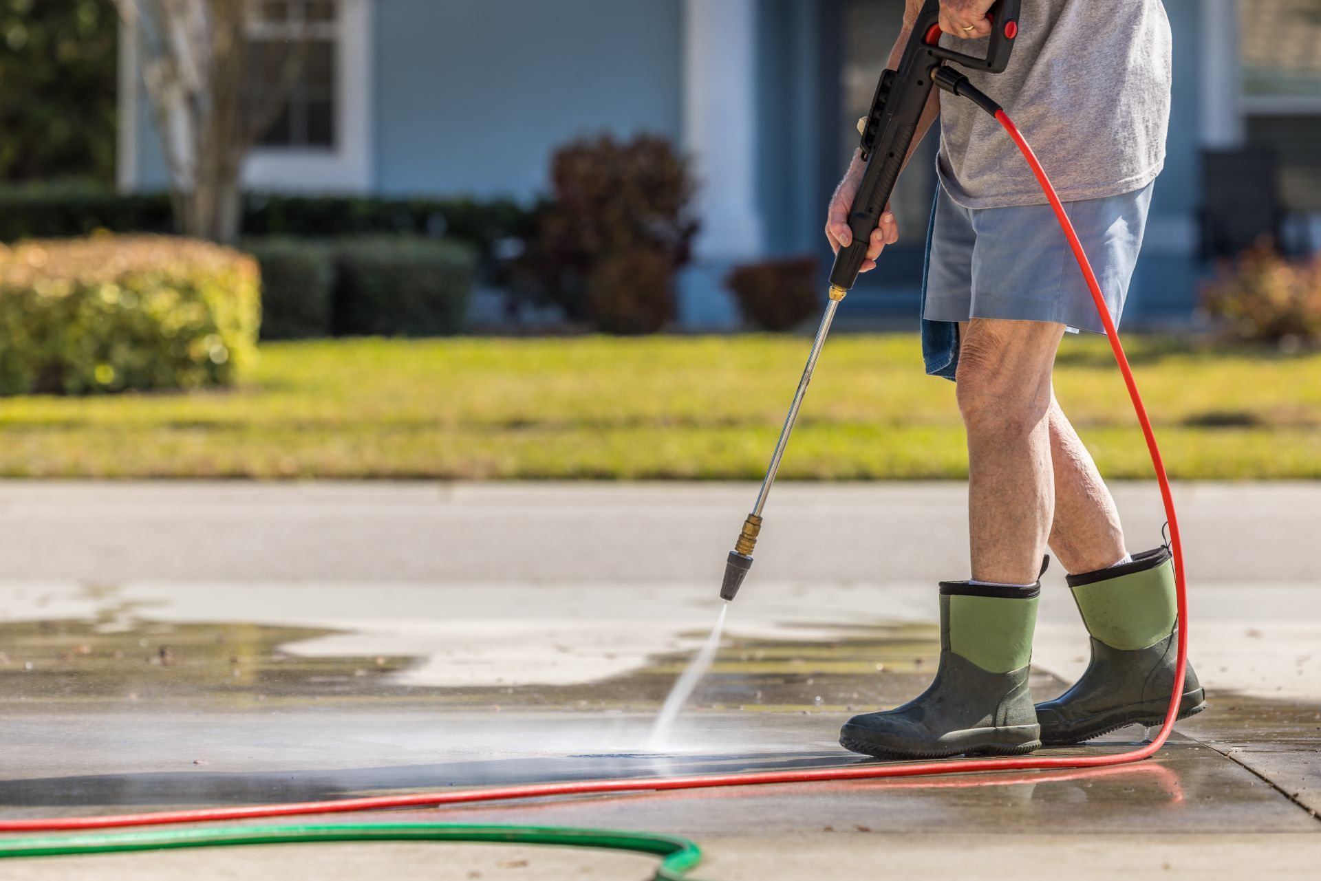 Person pressure-washing a driveway with a hose and spray wand, wearing green boots.