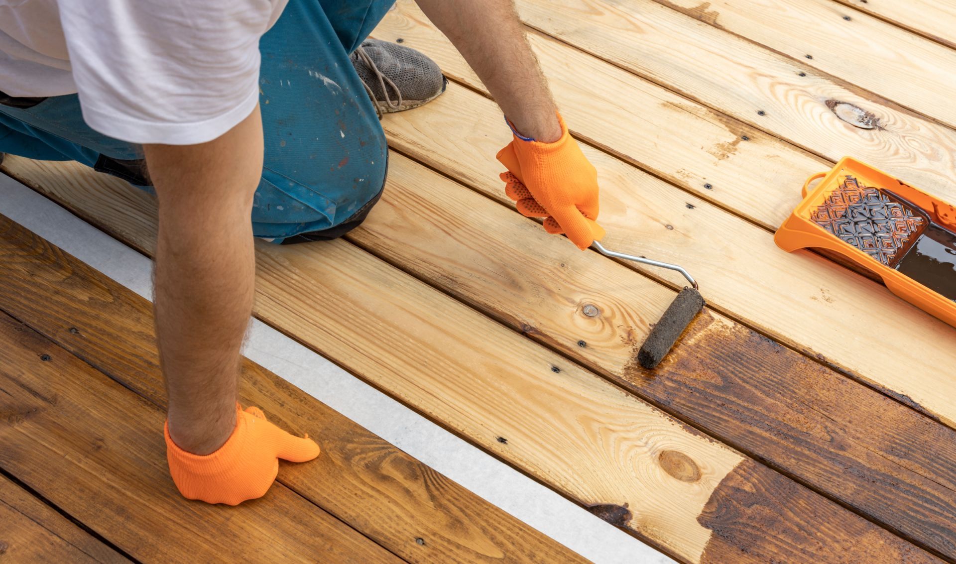 Worker staining wooden deck boards with a brush while wearing orange gloves
