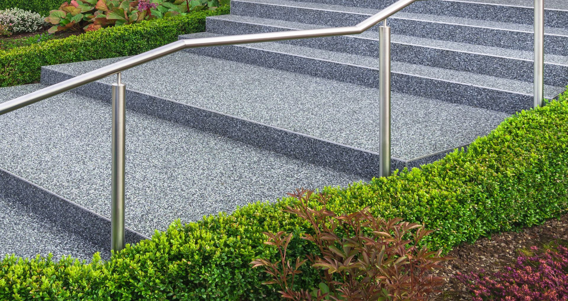 Gravel stairway with metal handrails beside a green landscaped slope and flowerbed