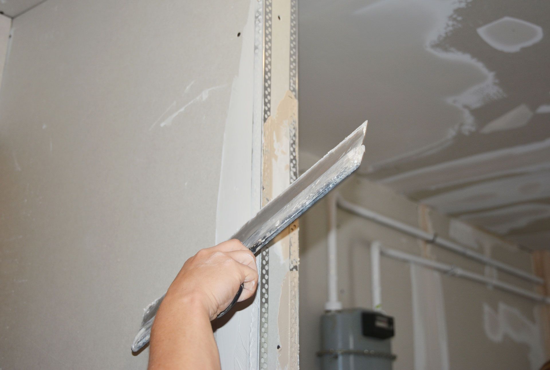 Hand smoothing drywall compound on a corner seam with a metal taping knife in an unfinished room
