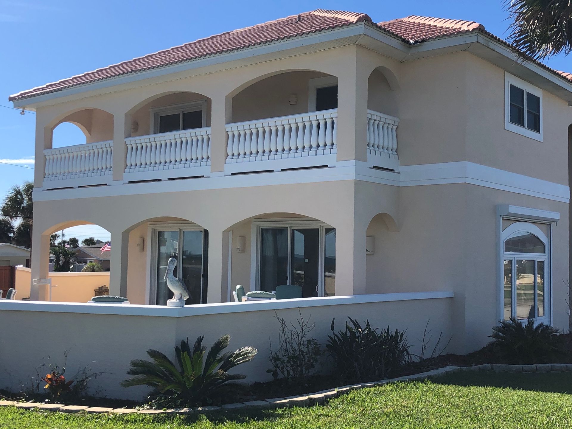 Large two-story beige house with red tile roof, white balconies, palm trees, and green lawn under a blue sky
