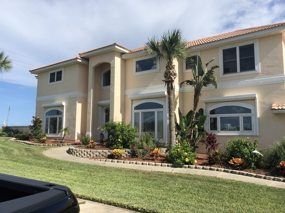 Two-story beige stucco house with arched windows, palm trees, and a landscaped front lawn under a blue sky