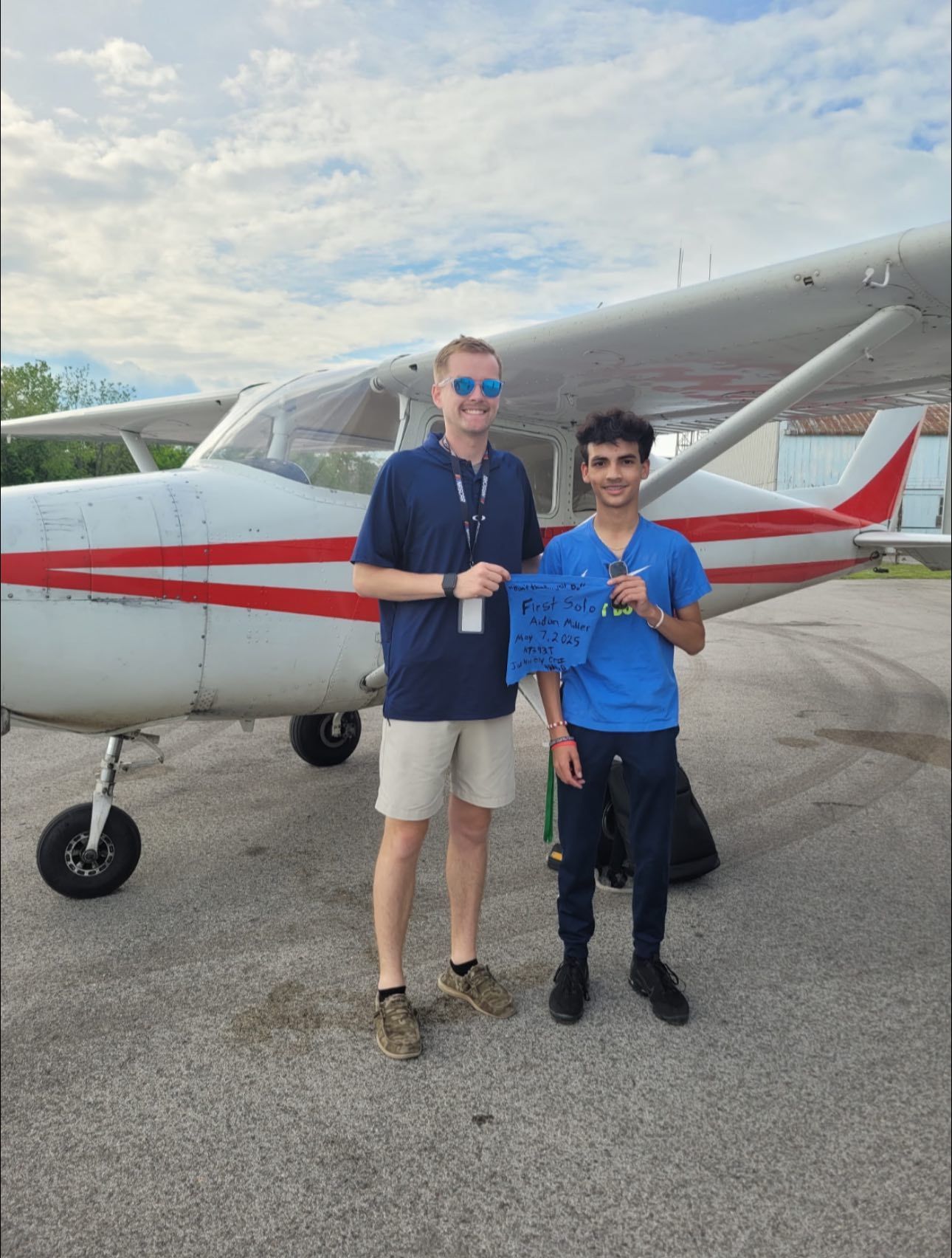 Two men standing next to a small airplane. One holds a blue paper. Sky in the background.