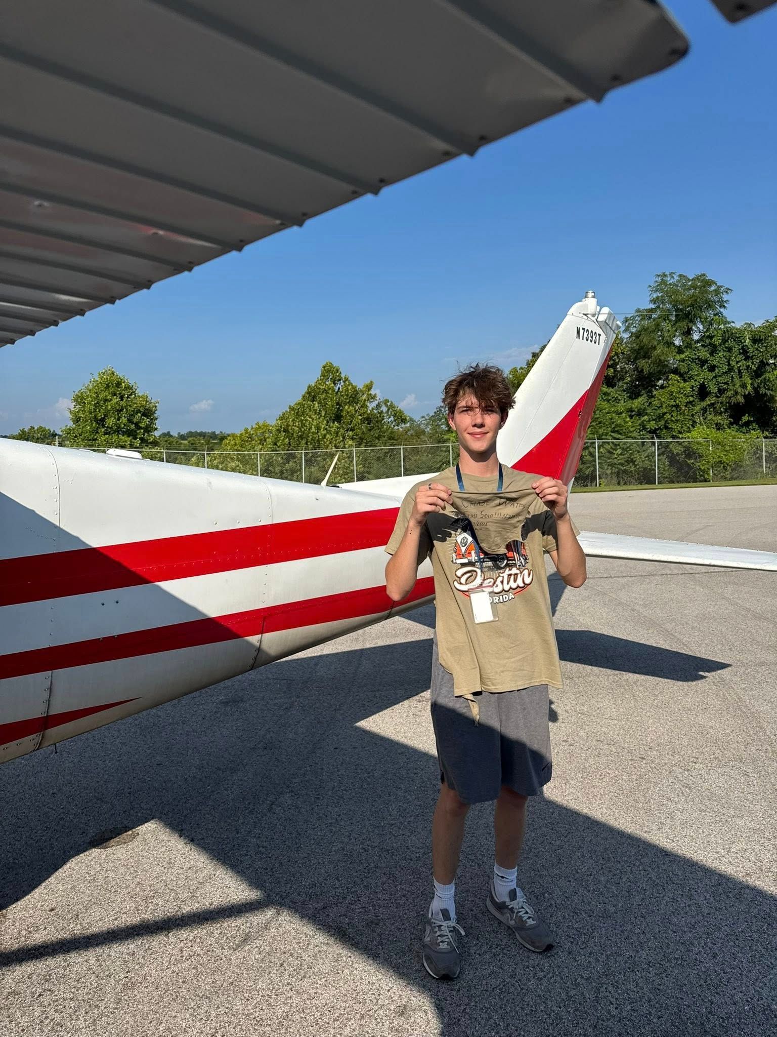 Teenager holding a shirt in front of a small airplane with red and white stripes on the tail.
