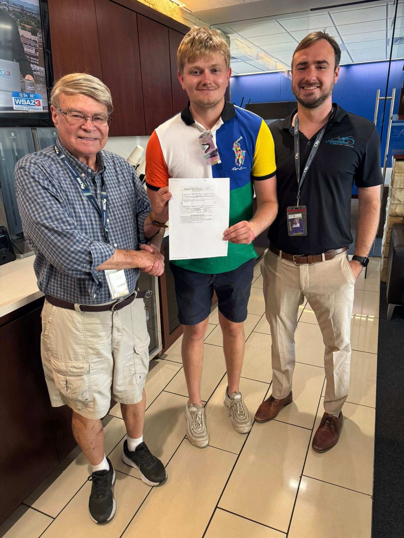 Three men stand indoors. One holds paper, wearing a polo. Others stand on either side.