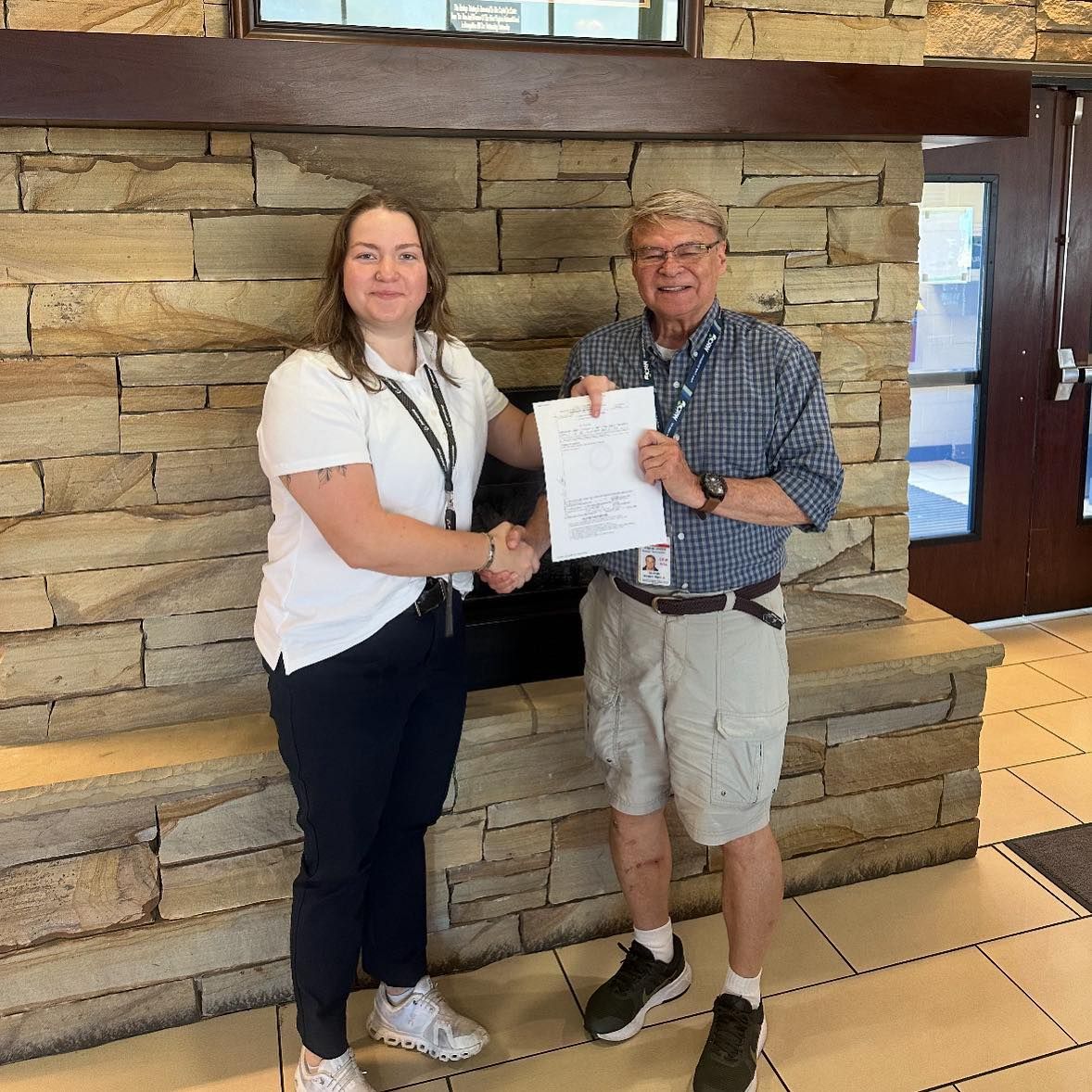 Woman and man shake hands, holding a document, in front of a stone fireplace.