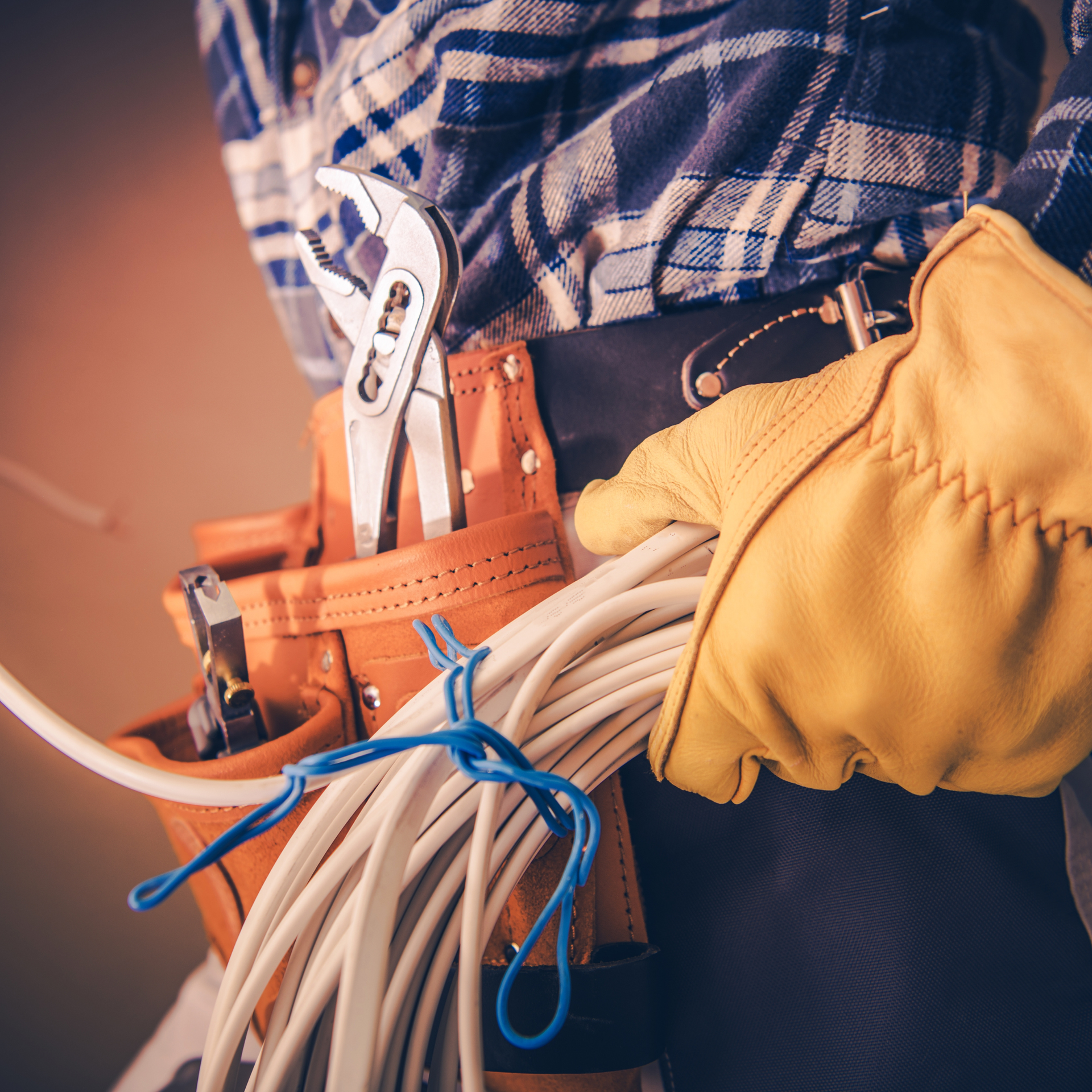 Electrician in plaid shirt, holding wire, wearing a tool belt and yellow glove.