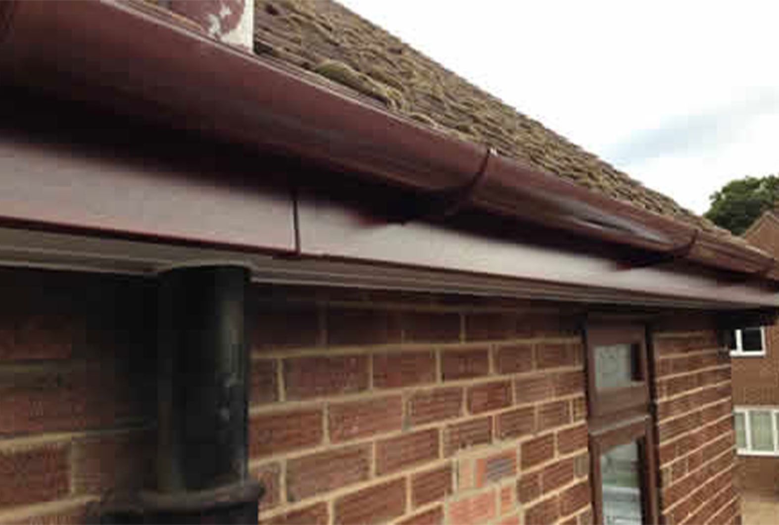 Brown gutters on a brick building with a dark downspout, against a roof with a cloudy sky.