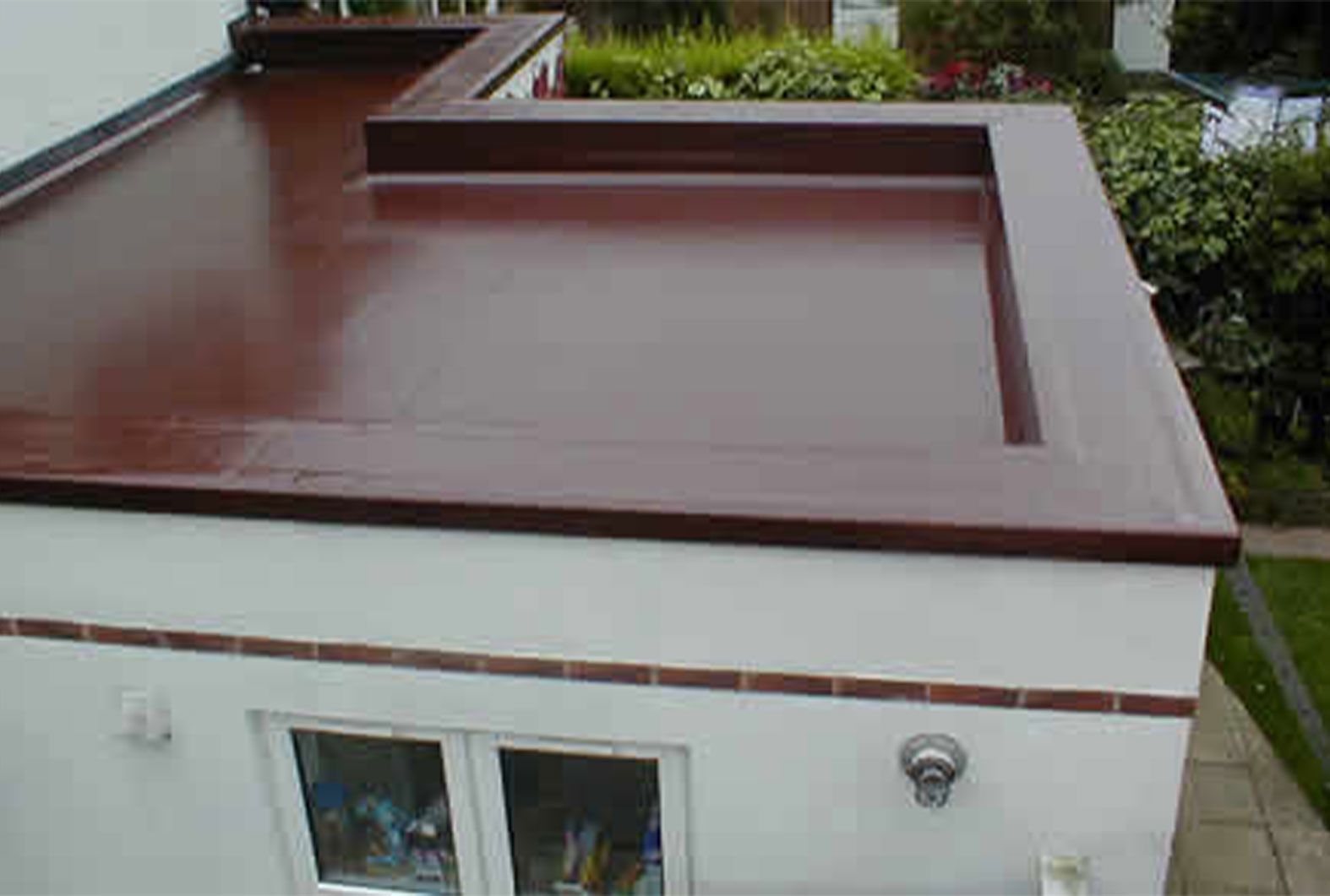 Brown flat roof on a white building with brick trim. Green plants in background.