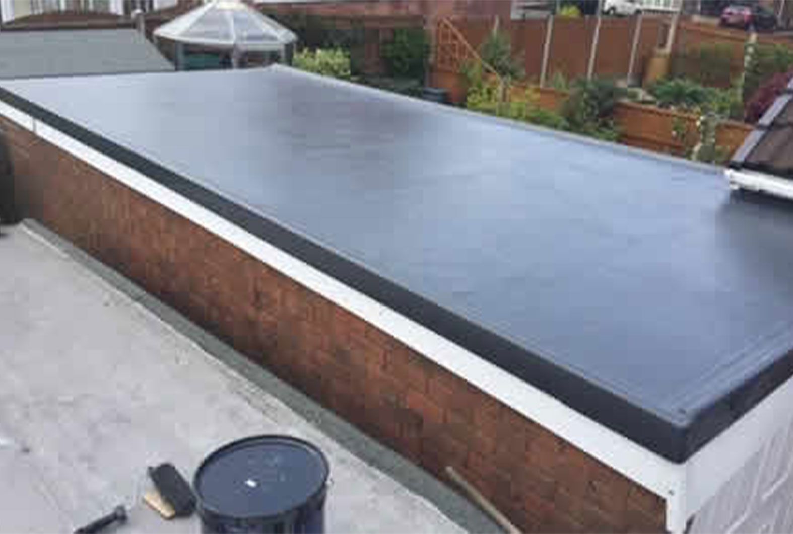 Black flat roof on a brick building with white trim, a garden in the background, and a bucket in the foreground.