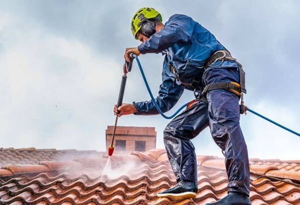 Man on a roof power washing it, wearing safety gear and a helmet.