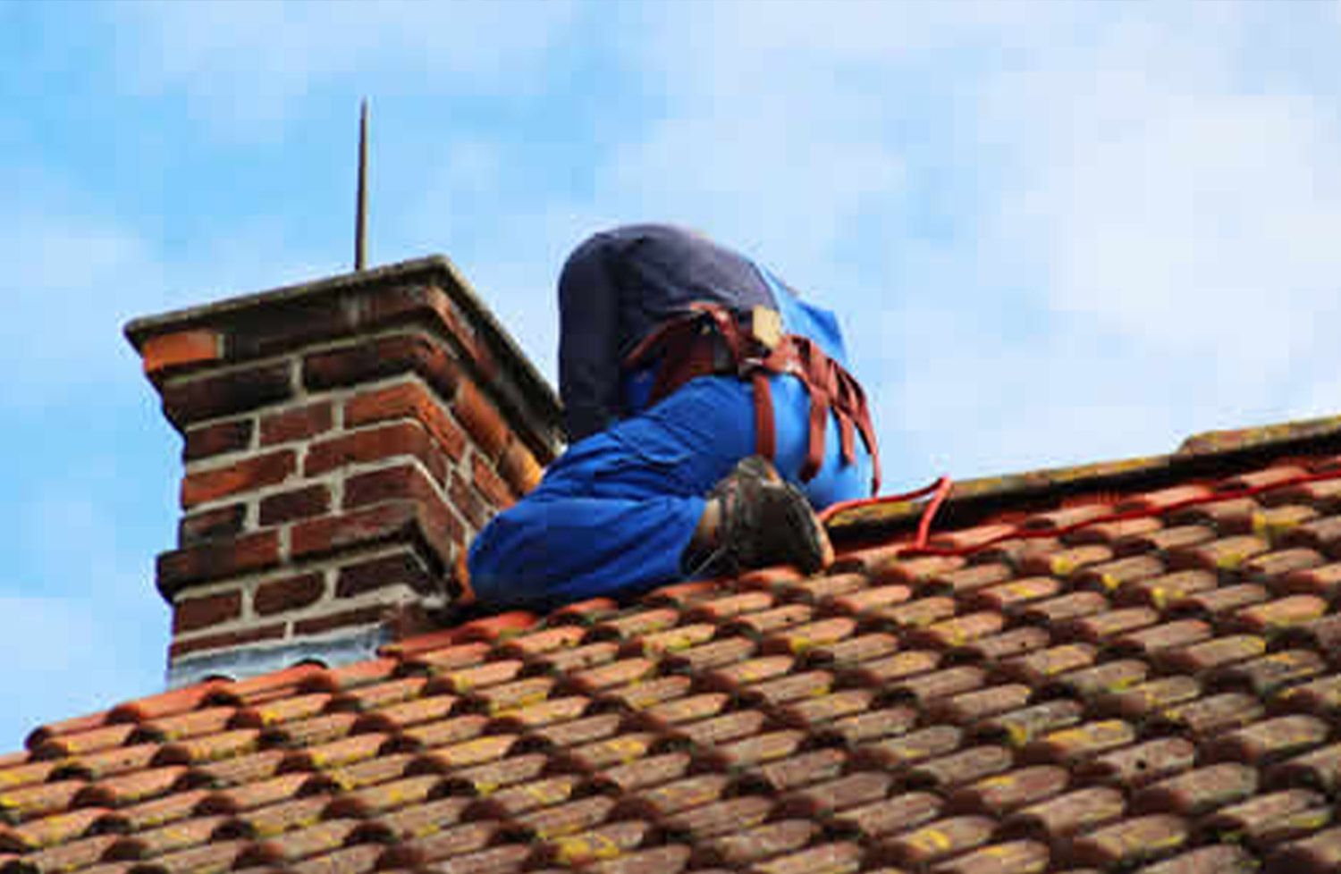 Person on a roof repairing a brick chimney; blue jumpsuit, safety harness, red-tiled roof, cloudy sky.