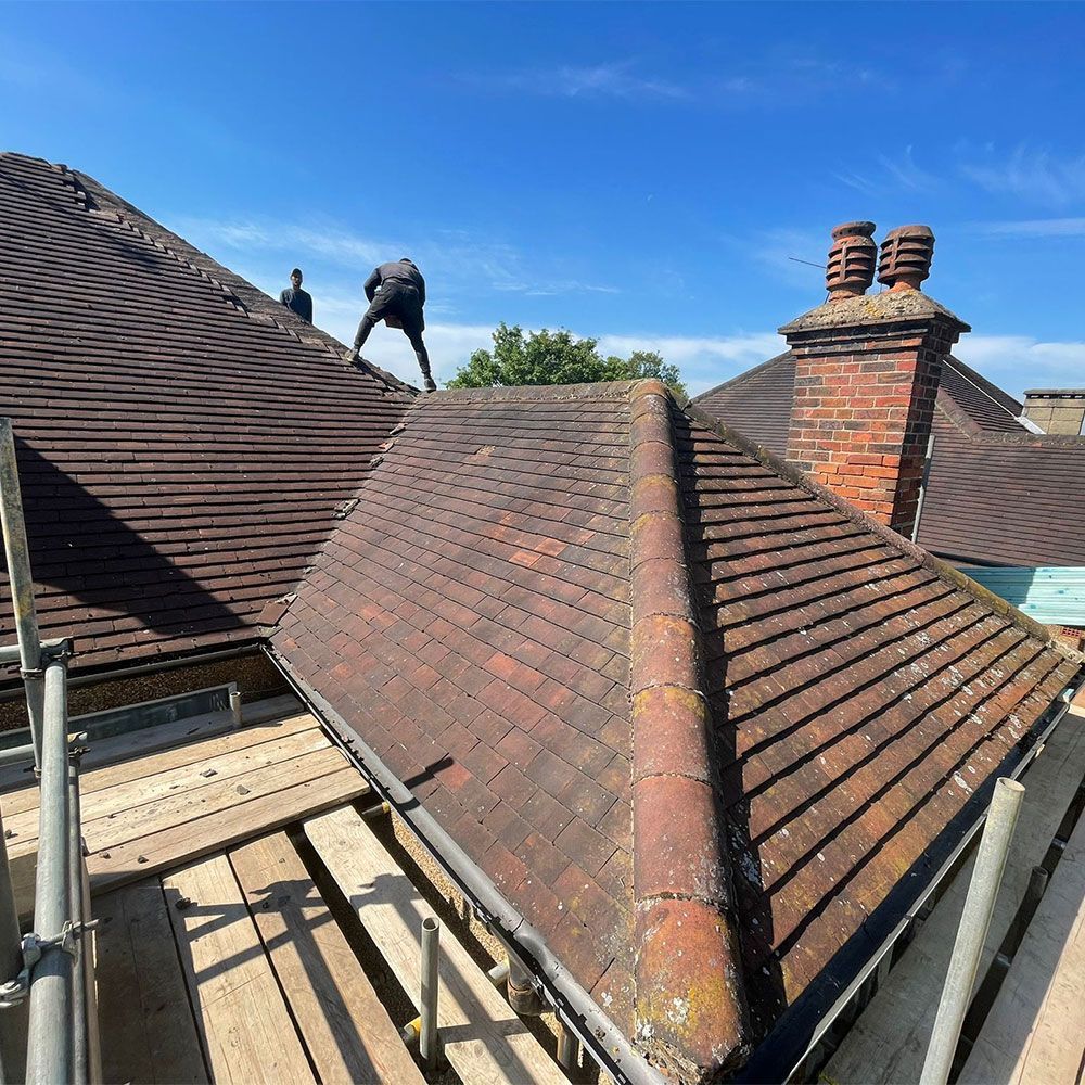 Workers on a rooftop with terracotta tiles, scaffolding, and brick chimney against a blue sky.