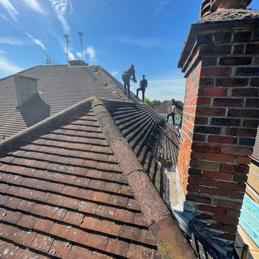 Three people on a tiled roof near a brick chimney on a sunny day.